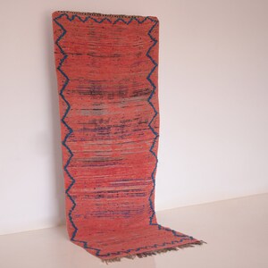 May include: A rectangular runner rug featuring a vibrant coral-red base with horizontal streaks of dark blue and black. The rug is framed by a prominent dark blue zigzag border pattern that runs along both long sides and the bottom edge.