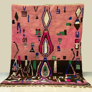Peut inclure: Tapis marocain rose et marron avec des motifs géométriques. Le tapis présente une forme de losange central avec des accents bleus, blancs et roses. Diverses formes et symboles bleus, verts et noirs sont dispersés sur le fond rose.