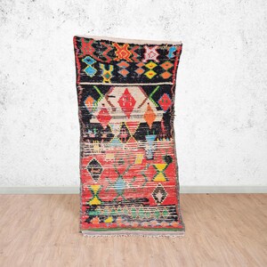 Peut inclure: Un tapis rectangulaire tissé à la main avec un design géométrique vibrant. Le tapis présente un fond rouge et noir avec des motifs de losanges et de croix colorés en jaune, bleu, orange et blanc. Le tapis est présenté contre un mur neutre et un sol en bois.