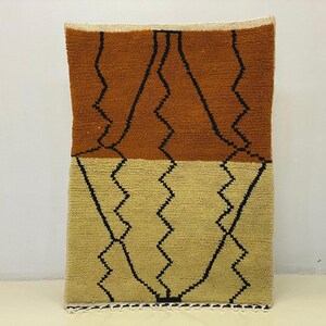 Peut inclure: Un tapis rectangulaire tissé à la main avec un design bicolore. La partie supérieure est marron chaud, tandis que le bas est beige clair. Des motifs en zigzag noirs courent verticalement. Le tapis a un aspect texturé et moelleux.