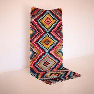 Peut inclure: Un tapis marocain coloré avec un motif géométrique en forme de losange dans des tons de rouge, bleu, jaune, vert et blanc. Le tapis a une bordure frangée.