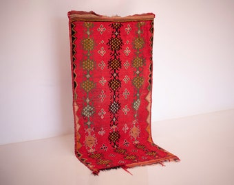 May include: A vibrant red rug with geometric patterns in black, green, and yellow. The rug has a central vertical design with diamond and lozenge shapes. The edges are trimmed with a black border. The rug is displayed against a white background.