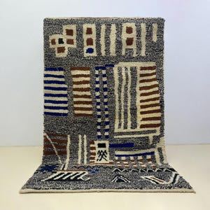 Peut inclure: Un tapis rectangulaire avec un fond gris et un motif géométrique. Le design comprend des blocs et des rayures dans des tons de bleu, de marron et de crème. Le tapis est fait d'une matière texturée et a un aspect doux et moelleux.