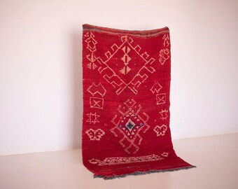 May include: A vibrant red rug with geometric patterns in white and accents of blue and gold. The rug features a central diamond design and decorative borders. The rug is displayed against a white wall.