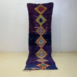 May include: A rectangular, hand-woven rug with a deep purple background and geometric diamond patterns in various colors. The rug features a dark blue section and is approximately 6 feet long.