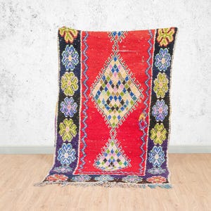 Peut inclure: Un tapis rectangulaire vibrant avec un champ central rouge et un motif en losange au centre. Le tapis présente une bordure noire avec des motifs floraux bleus, verts et violets. Le tapis est en matière tissée.