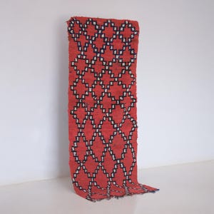 May include: A rectangular woven rug featuring a vibrant red base color with a repeating geometric diamond pattern outlined in black and white. The textile has a textured surface and a consistent grid-like design that spans the entire length.