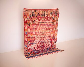 Puede incluir: Una alfombra tejida roja, naranja y azul con un patrón geométrico. La alfombra tiene flecos en la parte inferior.
