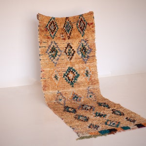 May include: A tan and brown woven rug with a diamond pattern in teal, orange, and blue. The rug has a textured, fringed edge and is leaning against a white wall. The rug is a decorative floor covering.