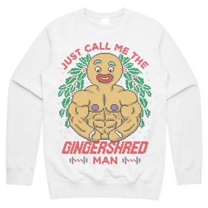 May include: White sweatshirt featuring a pixelated gingerbread man with a muscular physique. The text "JUST CALL ME THE GINGERSHRED MAN" is printed above and below the gingerbread man. The design includes holly and weights.