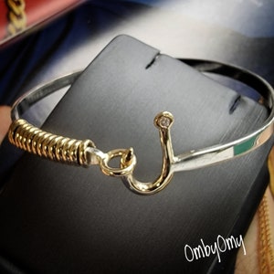 May include: A silver bangle bracelet with a gold hook clasp and a single diamond accent. The hook is shaped like a fish hook.