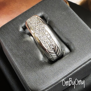 Puede incluir: Un anillo de plata con una parte superior rectangular incrustada con pequeñas piedras brillantes. El anillo tiene un diseño texturizado en los lados y se presenta en una caja de joyería negra. Es una joya.