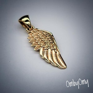 Puede incluir: Un colgante dorado con forma de ala estilizada. El ala presenta detalles de plumas y un lazo para una cadena. El colgante está sobre un fondo suave y neutro. El texto "OmbyOmy" está en la esquina inferior derecha.