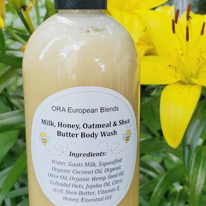 May include: A white plastic bottle with a black lid and a white label. The label says "ORA European Blends Milk, Honey, Oatmeal & Shea Butter Body Wash" and lists the ingredients. The bottle is in front of a yellow flower.
