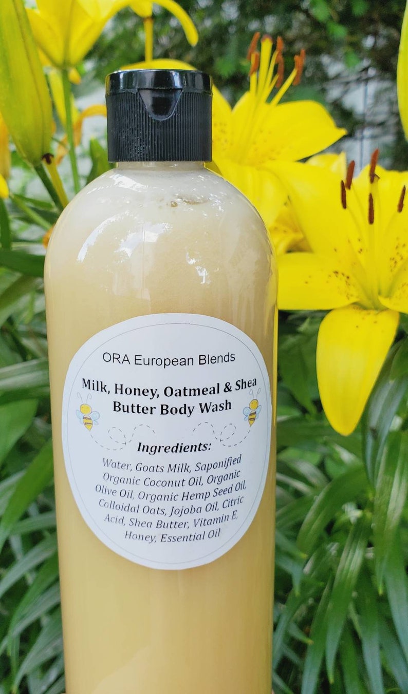 May include: A white plastic bottle with a black lid and a white label. The label says "ORA European Blends Milk, Honey, Oatmeal & Shea Butter Body Wash" and lists the ingredients. The bottle is in front of a yellow flower.