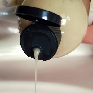 May include: A clear plastic bottle with a black cap is being squeezed, dispensing a white liquid.