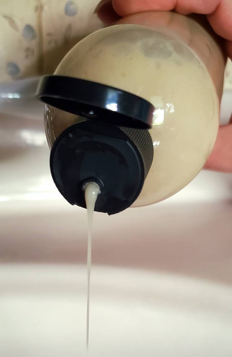 May include: A clear plastic bottle with a black cap is being squeezed, dispensing a white liquid.