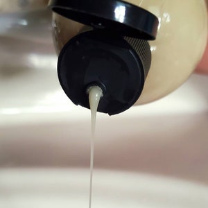 May include: A white liquid is being poured from a beige bottle with a black cap. The liquid is forming a thin stream.