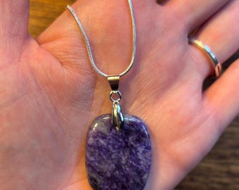 May include: A silver chain necklace with a rectangular, purple pendant. The pendant has a marbled appearance with shades of purple and gray. The necklace is displayed on a hand.