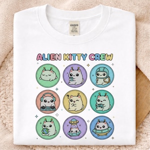 May include: White t-shirt featuring the text "ALIEN KITTY CREW" above nine circular illustrations of cartoon cats with alien features. Each cat is in a different pose, with various accessories and backgrounds. The design is in pastel colors.
