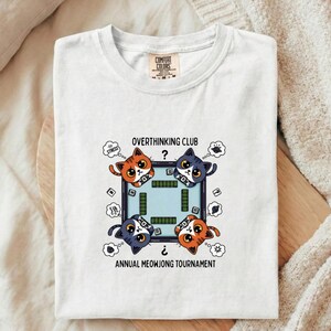 May include: White t-shirt with a graphic of four cartoon cats playing mahjong. Text reads "OVERTHINKING CLUB?" and "ANNUAL MEOWJONG TOURNAMENT". The cats are orange and blue, with thought bubbles and stress symbols.