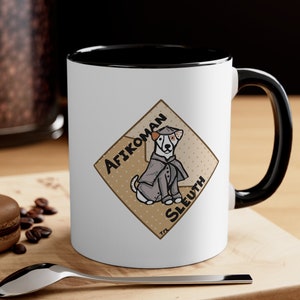 May include: White ceramic mug with a black interior and handle. The mug features a cartoon dog wearing a detective's outfit, with the words "Afikoman Sleuth" on a diamond-shaped background.