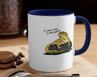 Puede incluir: Taza de cerámica blanca con borde y asa azules. La taza presenta una imagen de dibujos animados de un dragón barbudo sentado en una roca con el texto "Soy fuego, soy muerte."