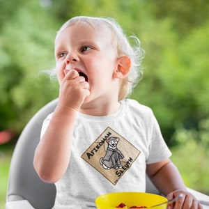 May include: A toddler wearing a white t-shirt with a graphic of a dog detective in a diamond-shaped design. The text on the design reads "Afikoman Sleuth". The child is sitting in a high chair and eating from a yellow bowl.