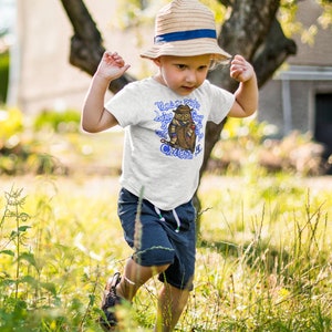 May include: A young child wearing a white t-shirt with a blue owl graphic and the text "That's no point in being grumpy if you can't criticize it". The child is wearing a straw hat with a blue band and blue shorts. The child is standing in a grassy field.