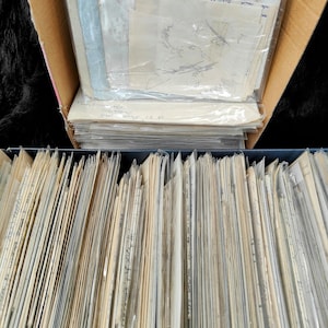 May include: A close-up view of a large stack of old, yellowed paper documents stored in a cardboard box and a black file box. The documents are organized and stacked neatly.