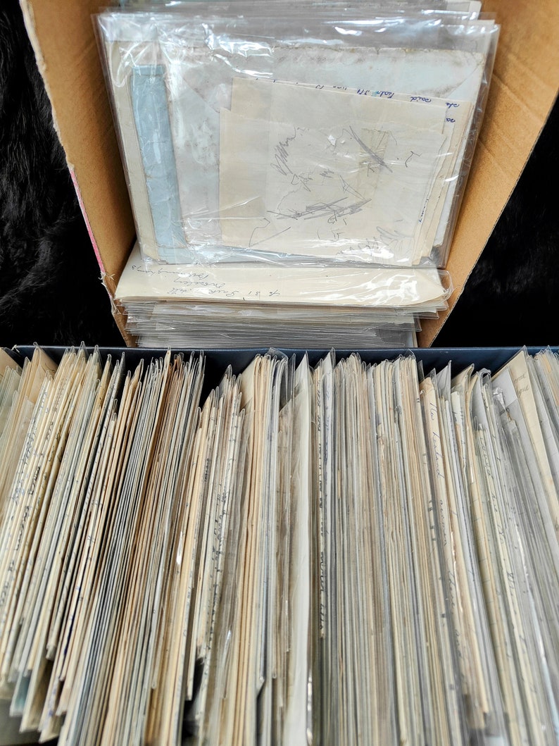May include: A close-up view of a large stack of old, yellowed paper documents stored in a cardboard box and a black file box. The documents are organized and stacked neatly.