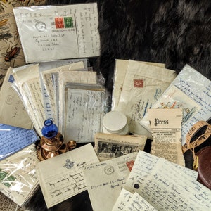 May include: A collection of vintage letters, envelopes, and postcards, some with stamps, are piled on a brown fur surface. There is a small blue glass bottle, a copper candle holder, a white jar, a brown leather case with a metal clasp, and a newspaper with the word "Press" on the front.