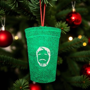 May include: A green Christmas tree ornament shaped like a coffee cup, with a white graphic of a face. The ornament is hanging from a red string against a backdrop of a Christmas tree with red ornaments.