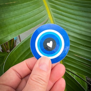 May include: A round sticker featuring a blue and white evil eye design with a white heart in the center. The sticker is held in a hand against a backdrop of green leaves.