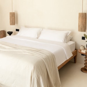 May include: A white bed with a white linen blanket and two white pillows. The bed is in a bedroom with a wooden cabinet and a side table with a plant and a vase.