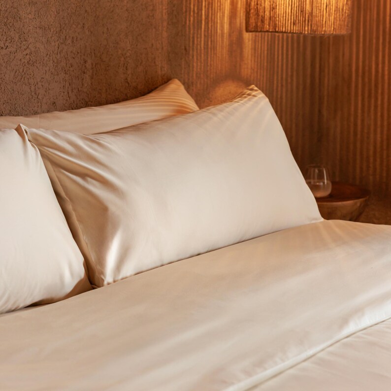 May include: Beige satin bed linens, including a pillow and a duvet cover, are arranged on a bed. The pillowcases have a subtle striped pattern. The scene is illuminated by warm, ambient lighting, creating a cozy atmosphere. The bedding is smooth and wrinkle-free.