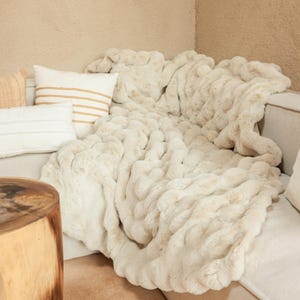 May include: A soft, cream-colored faux fur throw blanket draped over a couch with pillows. The blanket has a wavy, textured pattern.