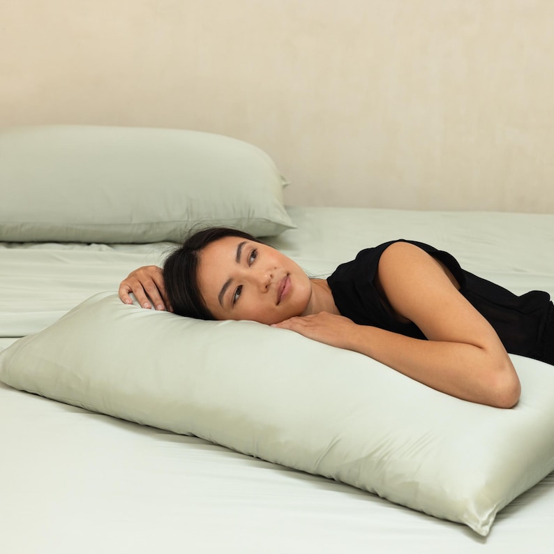 May include: A woman is lying on a bed with a light green pillow and a light green sheet. She is wearing a black sleeveless top and has her head resting on the pillow.