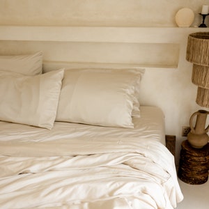 May include: A white bed with a white duvet cover and two white pillows. The bed is made and the duvet is folded back. The bed is in a room with white walls and a shelf above the headboard.