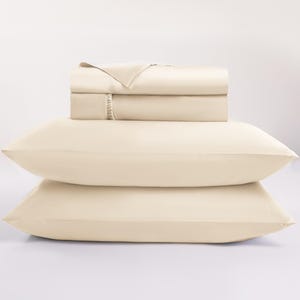 May include: A cream-colored bed sheet set, including two pillowcases and a folded sheet set. The sheets are stacked neatly, with a smooth, soft texture. The pillowcases are positioned in front of the sheets, creating a layered effect. The bedding is a neutral color.