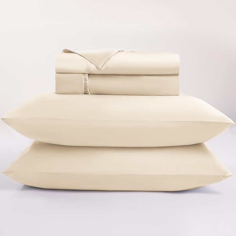 May include: A cream-colored bed sheet set, including two pillowcases and a folded sheet set. The sheets are stacked neatly, with a smooth, soft texture. The pillowcases are positioned in front of the sheets, creating a layered effect. The bedding is a neutral color.