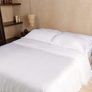 May include: A bed with white bedding, including a fitted sheet, flat sheet, and two pillows. The sheets and pillowcases have a scalloped edge with a subtle tan trim. The bed is in a neutral-toned room with wooden shelves and a hanging light fixture.