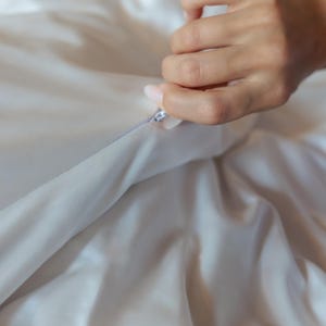 K&ouml;nnte beinhalten: Nahaufnahme eines wei&szlig;en Satin-Stoffs, der ge&ouml;ffnet wird. Der Stoff ist glatt und scheint ein Bettw&auml;scheartikel zu sein, wie ein Bettbezug oder ein Kissenbezug. Der Rei&szlig;verschluss ist silberfarben und der Stoff hat eine neutrale Farbe.