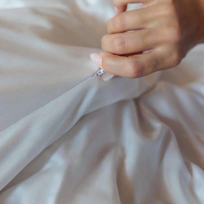 K&ouml;nnte beinhalten: Nahaufnahme eines wei&szlig;en Satin-Stoffs, der ge&ouml;ffnet wird. Der Stoff ist glatt und scheint ein Bettw&auml;scheartikel zu sein, wie ein Bettbezug oder ein Kissenbezug. Der Rei&szlig;verschluss ist silberfarben und der Stoff hat eine neutrale Farbe.