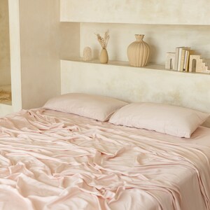 May include: A pink bed with a white headboard and two pink pillows. The bed is made with a soft, pink sheet and the fabric is wrinkled. The bed is in a room with a white wall and a shelf with decorative items.