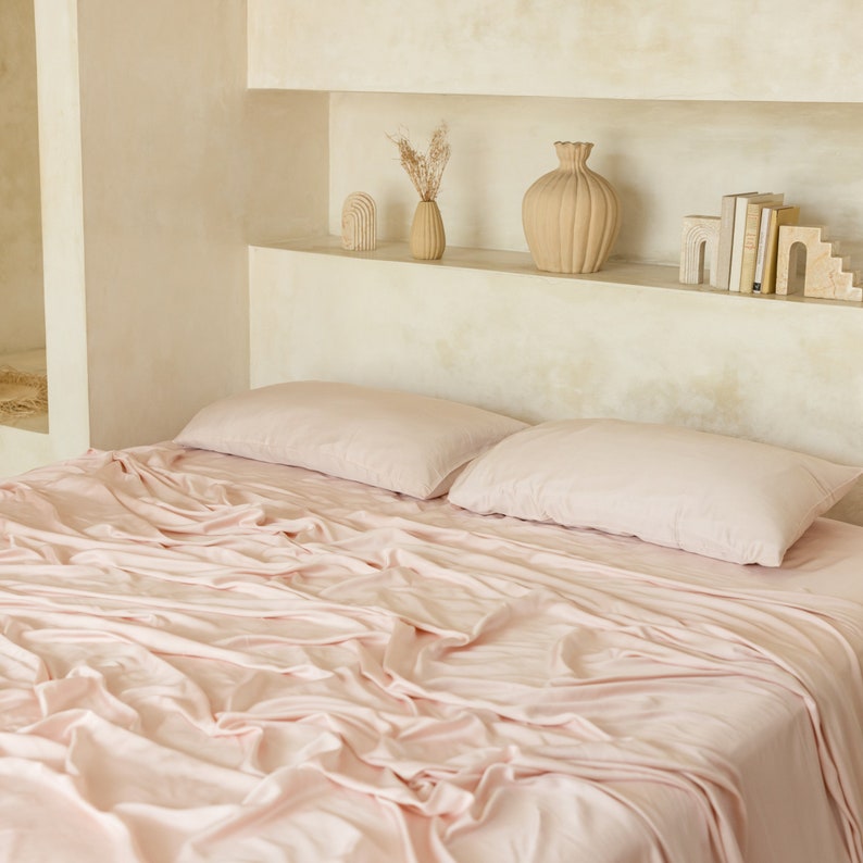 May include: A pink bed with a white headboard and two pink pillows. The bed is made with a soft, pink sheet and the fabric is wrinkled. The bed is in a room with a white wall and a shelf with decorative items.