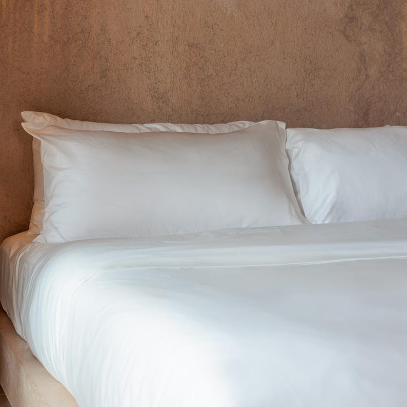 May include: A neatly made bed with crisp, white bedding. Two fluffy pillows rest against a textured, brown wall. The bed is covered with a smooth, white sheet and a matching duvet.