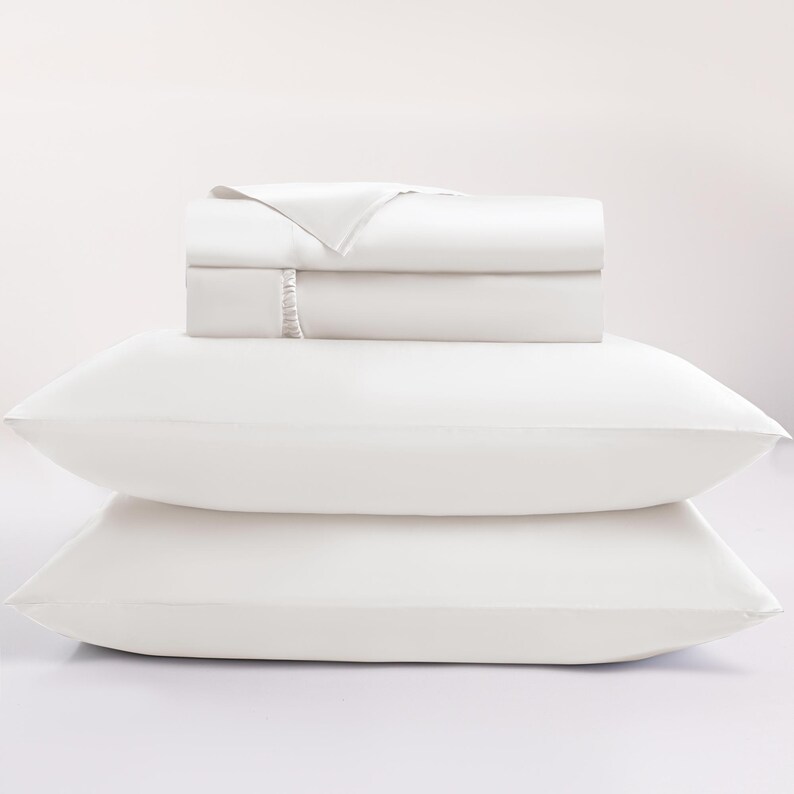 May include: A white bed sheet set with two pillows. The folded sheets are stacked on top of the pillows. The sheets and pillowcases are a solid white color. The image is well-lit and the background is white.