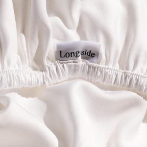 May include: Close-up of white fabric with elastic gathering. A small white tag with the words "Long side" in black lettering is sewn into the fabric. The fabric is smooth and appears to be a sheet or bedding.