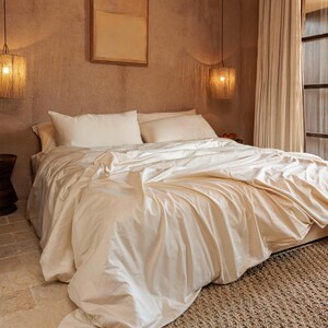 May include: A bed with cream-colored bedding, including a duvet cover and pillows. The bed is in a room with a neutral color palette, with a woven rug and a window with sheer curtains. Two pendant lights hang above the bed.